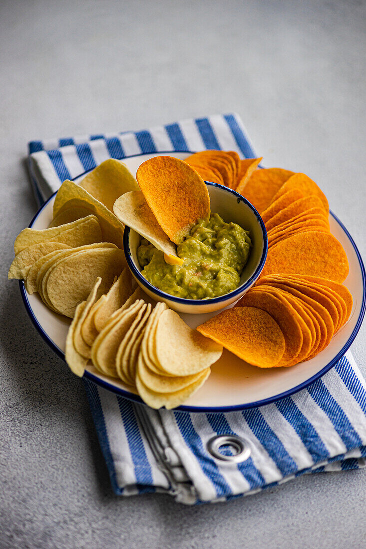 Top view of plate of assorted crisps including potato, paprika, and cheese flavors, served with a bowl of fresh avocado guacamole on a striped napkin