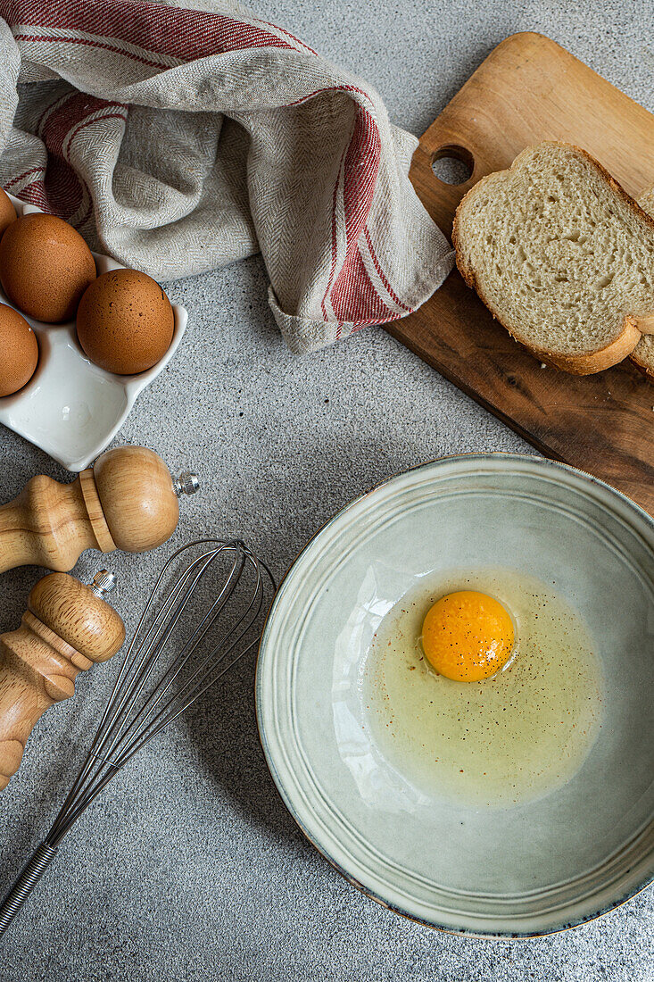 Top view of raw egg in bowl near cutting board with fresh bread while cooking at concrete table with various ingredients in kitchen