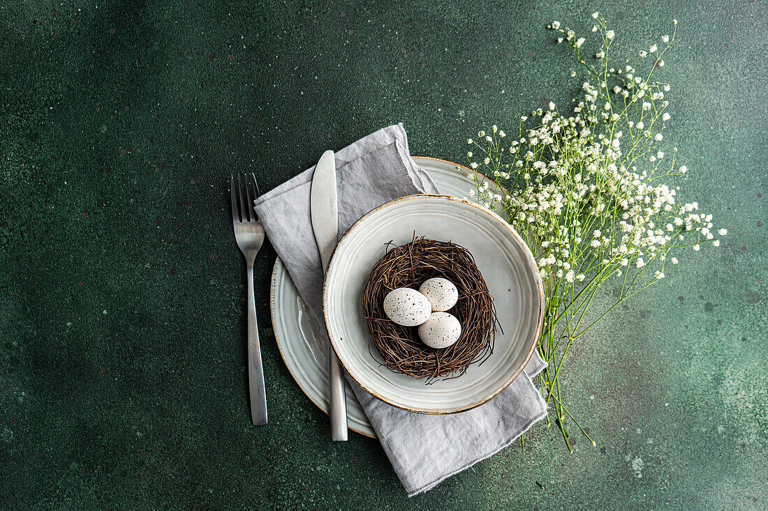 An artistic spring table setting featuring a bird's nest with speckled eggs, placed within layered crockery and accompanied by a delicate bouquet of baby's breath
