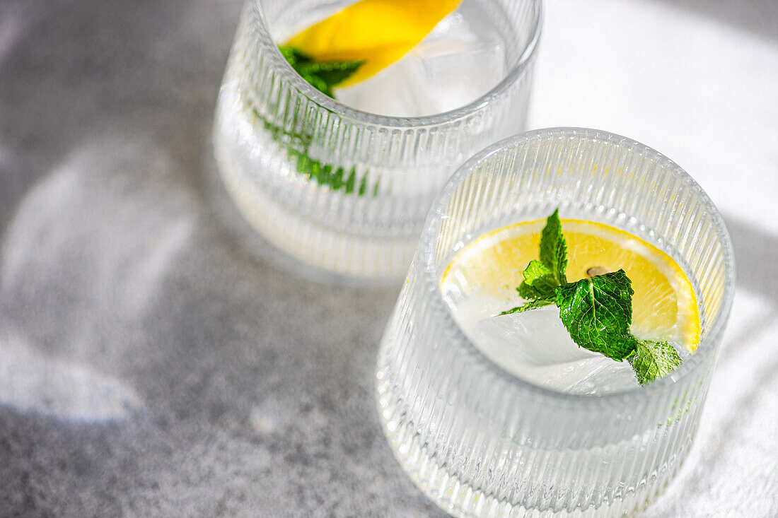 Two glasses of gin tonic with ice, garnished with fresh lemon slices and mint leaves, on a textured surface.