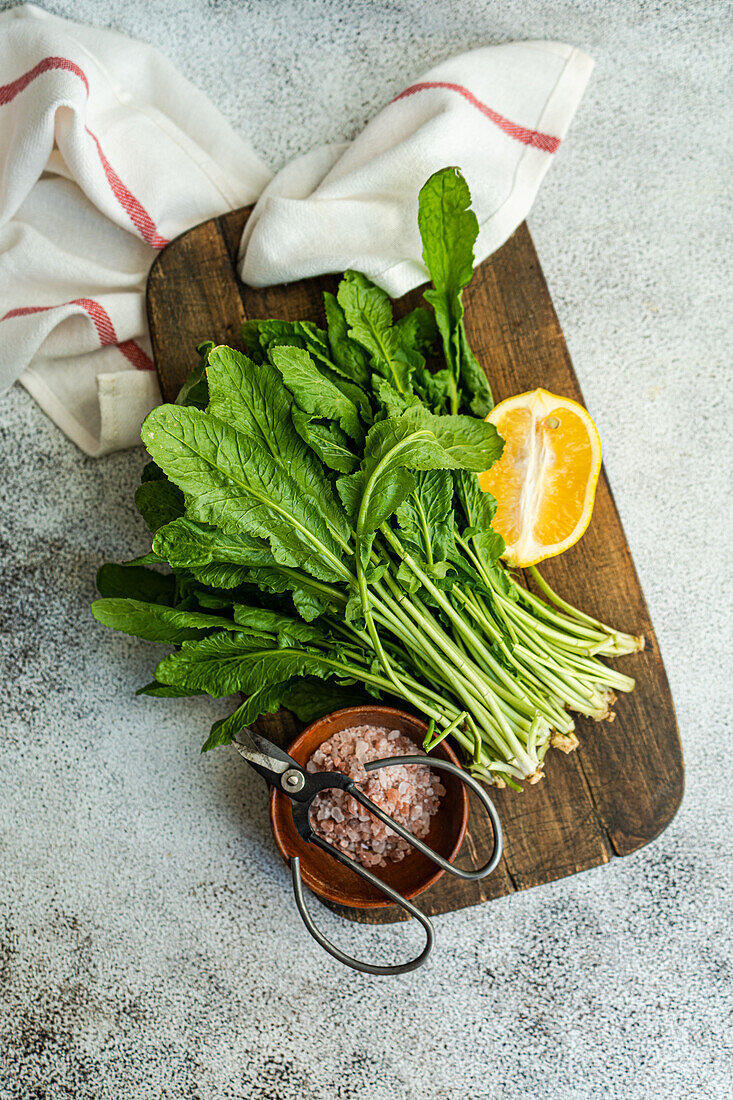 From above of fresh arugula leaves with a slice of lemon and a bowl of salt on a wooden cutting board, accented by kitchen scissors and a striped towel