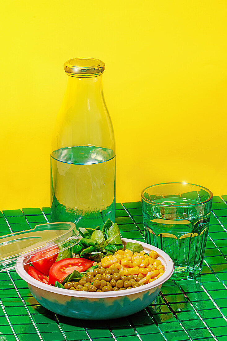Salatschüssel mit Tomatenscheiben, Spinatblättern, Maiskörnern und Erbsen auf grüner Fläche neben Flasche und Wasserglas vor gelber Wand