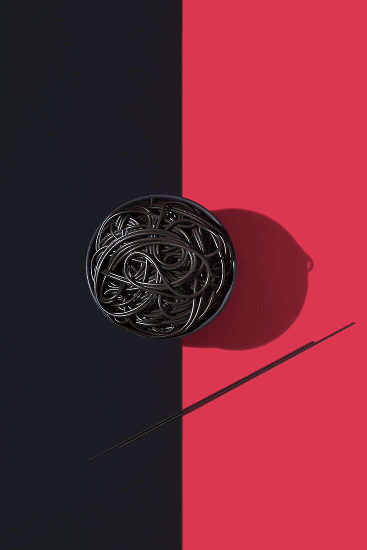 Draufsicht auf schwarze Spaghetti in einer Schüssel auf schwarzem und rotem Hintergrund neben Stäbchen