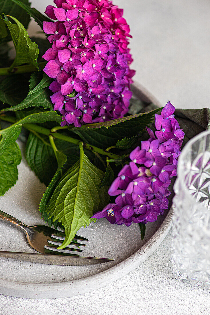 Draufsicht auf eine violette Hortensie, die auf einem weißen Tisch neben Keramiktellern und Gläsern steht