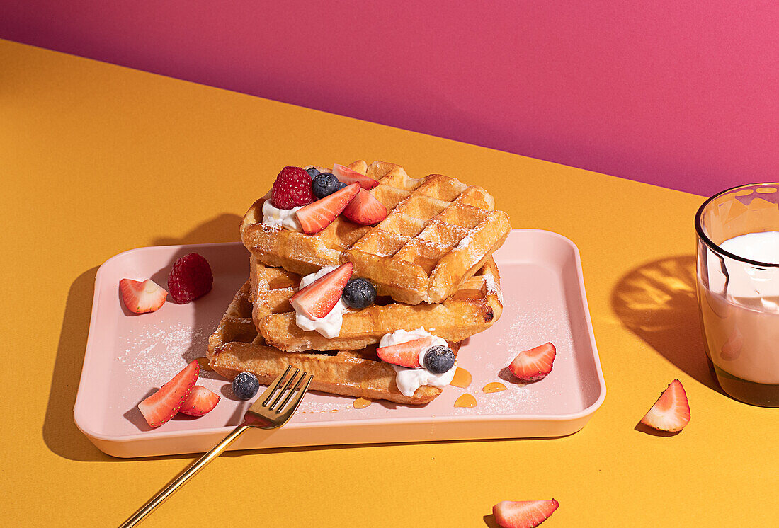 From above tasty sweet waffles topped with berries fruits sauce and cream served on pink tray on colorful table background near glass with hot milk beverage in light kitchen