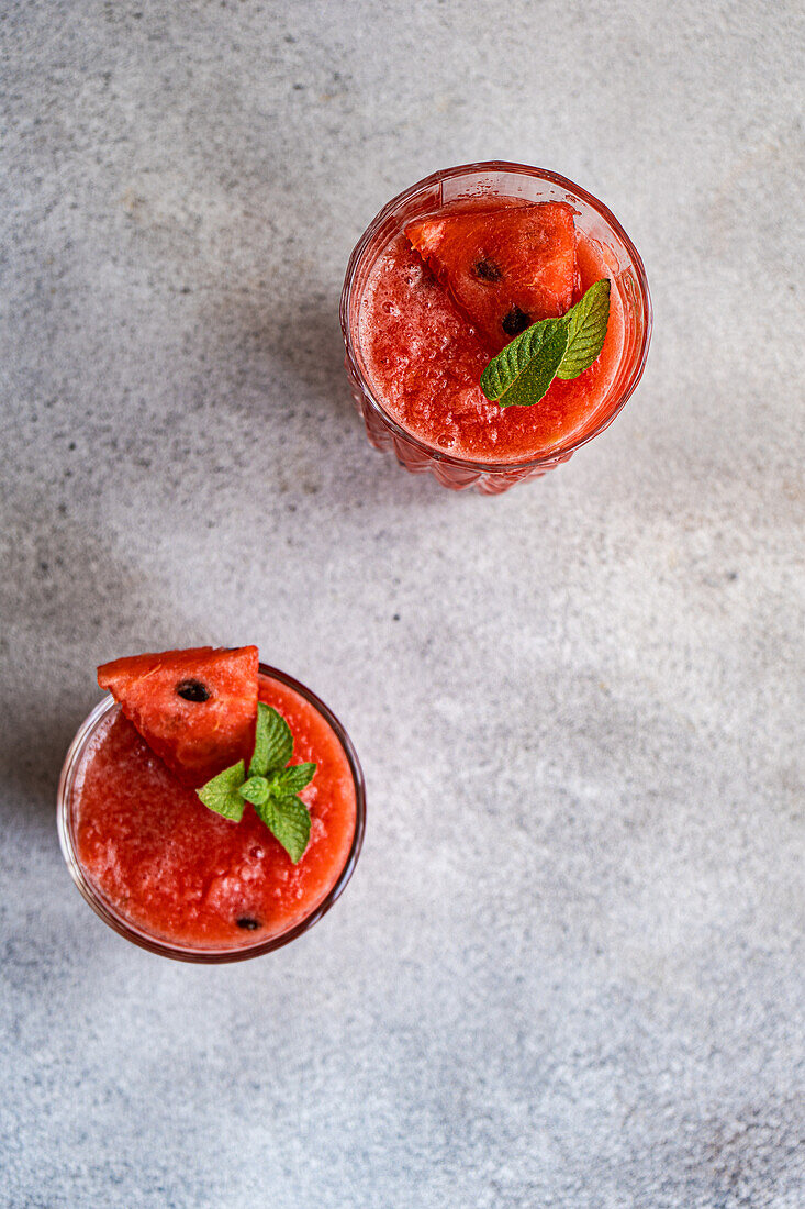 Top view of glasses of fresh Margarita cocktails with watermelon smoothieFrom above glasses of Margarita cocktail with watermelon smoothie garnished with leaves of mint placed on gray table covered with mint leaves