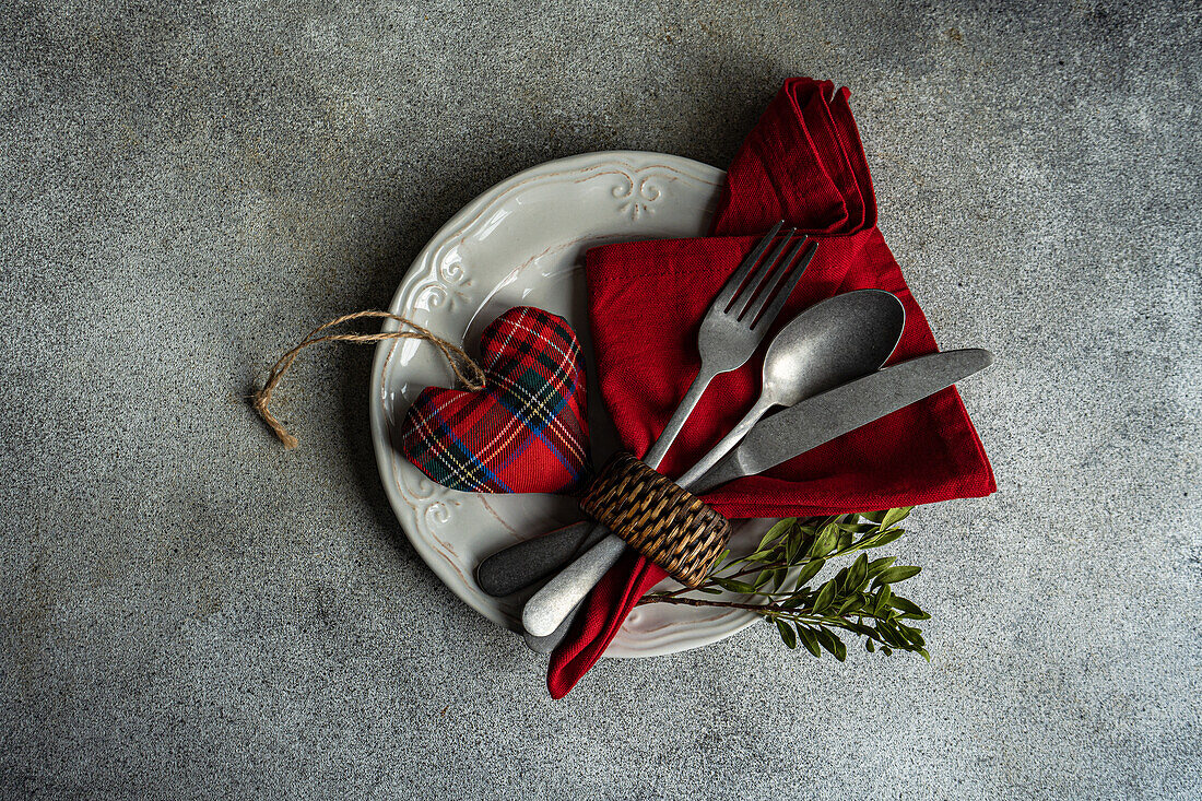 Draufsicht auf einen Keramikteller mit Besteck, eine Serviette aus Schottenstoff mit herzförmigem Dekor auf einer Betonfläche am Küchentisch für eine Mahlzeit während der Valentinstagsfeier
