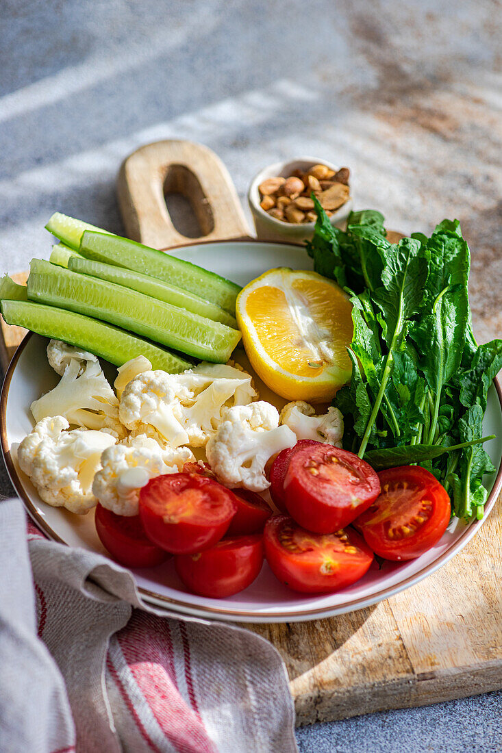 Eine gesunde Schüssel mit frischem Gemüse, darunter Gurkenscheiben, Kirschtomaten, Blumenkohlröschen, eine Zitronenspalte und Rucolablätter auf einem Holzbrett mit gemischten Nüssen