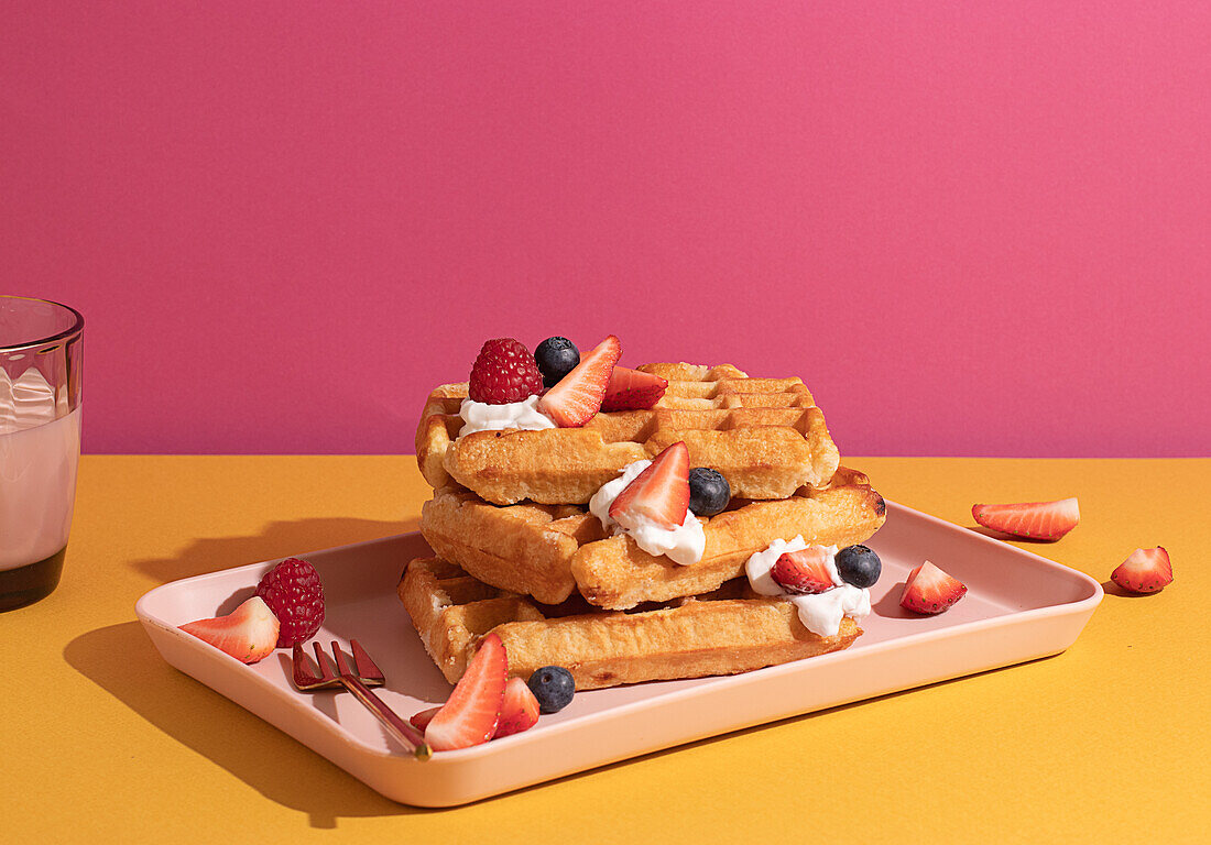 From above tasty sweet waffles topped with berries fruits sauce and cream served on pink tray on colorful table background near glass with hot milk beverage in light kitchen