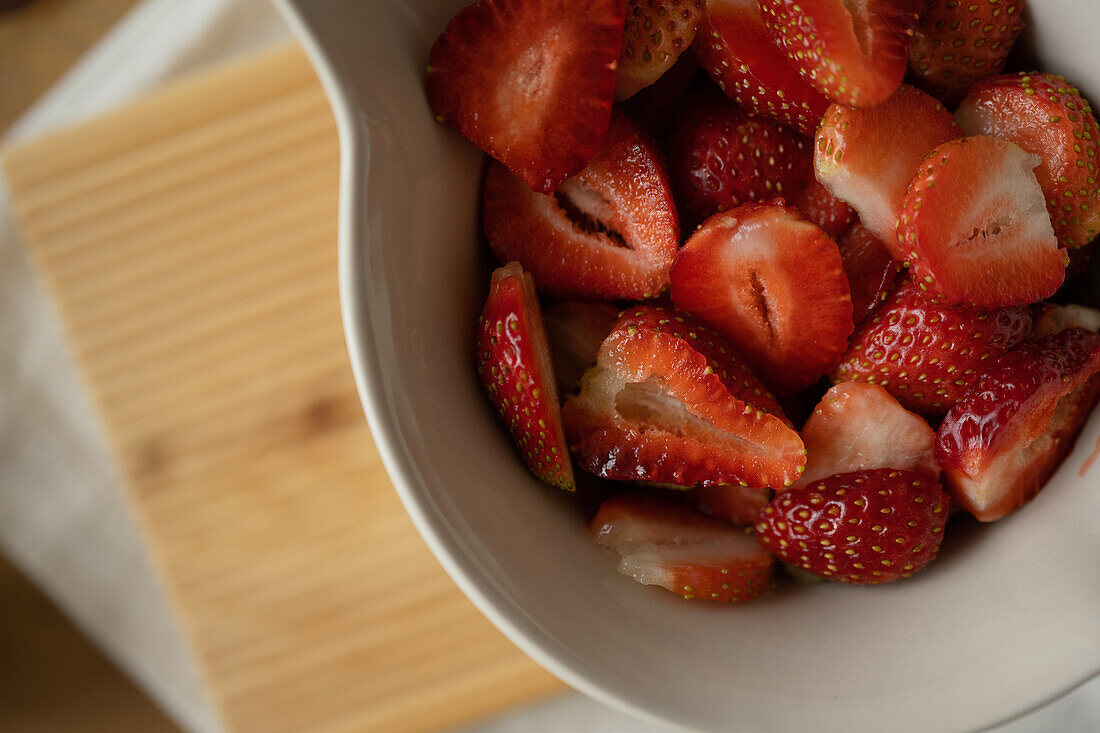 From above of appetizing sliced fresh strawberries on white ceramic plate on wooden board over table during preparation of jam