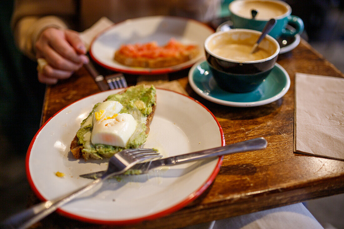 Leckerer knuspriger Toast mit Avocado und Ei auf einem Keramikteller mit Silberbesteck neben einer Tasse aromatischem Cappuccino in einer Cafeteria