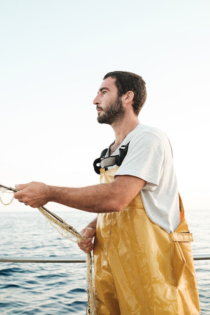 Seitenansicht eines bärtigen männlichen Fischers in Uniform, der mit seinem Netz auf Fischfang geht, während er auf einem Schoner in Soller nahe der Baleareninsel Mallorca arbeitet