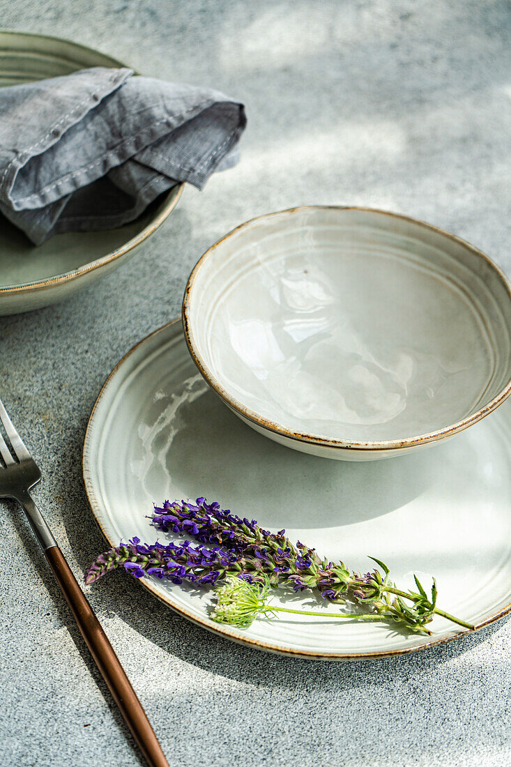 Hoher Winkel eines herbstlich gedeckten Tisches mit Keramikschüssel und -teller mit Lavendelblüten in der Nähe einer Gabel vor grauem Hintergrund