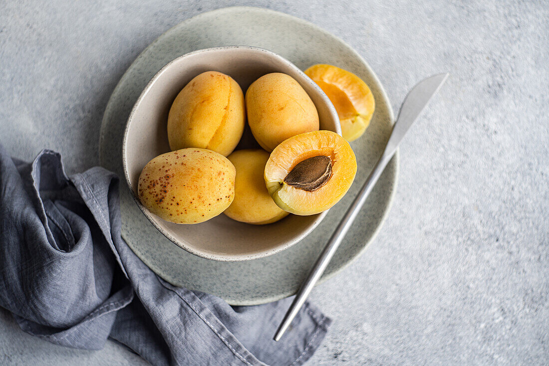 Top view of ceramic bowl with fresh ripe yellow apricots placed on blue cloth on concrete table with cut in half apricot