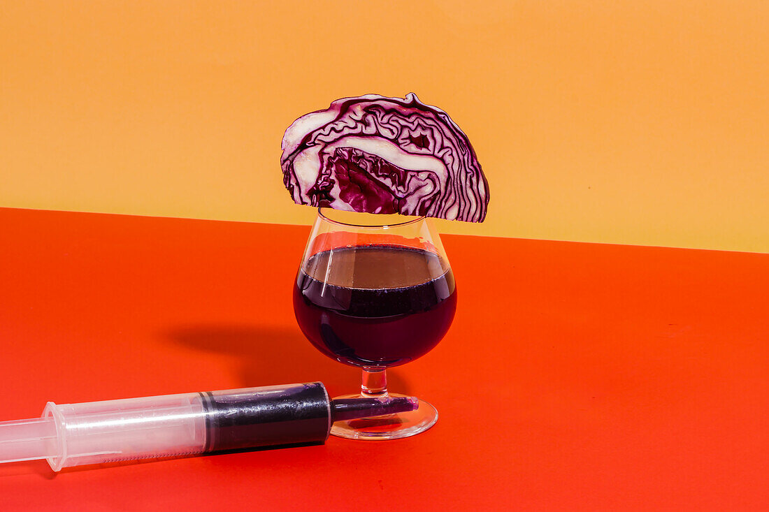 Komposition eines Glases mit hausgemachtem Rotkohlsaft mit Rotkohl darauf und neben einer Spritze vor orangefarbenem Hintergrund