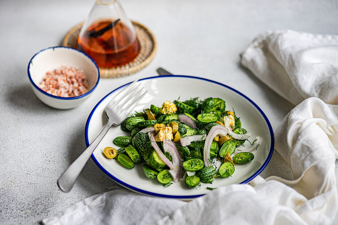 High angle of cucamelon salad with red onion, olives and spicy cheese served on white plate with fork and knife near napkin and oil on gray table in daylight