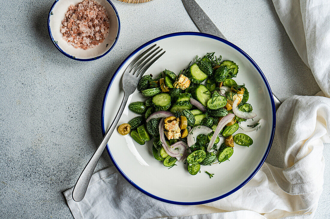 Draufsicht auf einen Gurkensalat mit roten Zwiebeln, Oliven und würzigem Käse, serviert auf einem weißen Teller mit Gabel und Messer neben einer Serviette auf einem grauen Tisch im Tageslicht