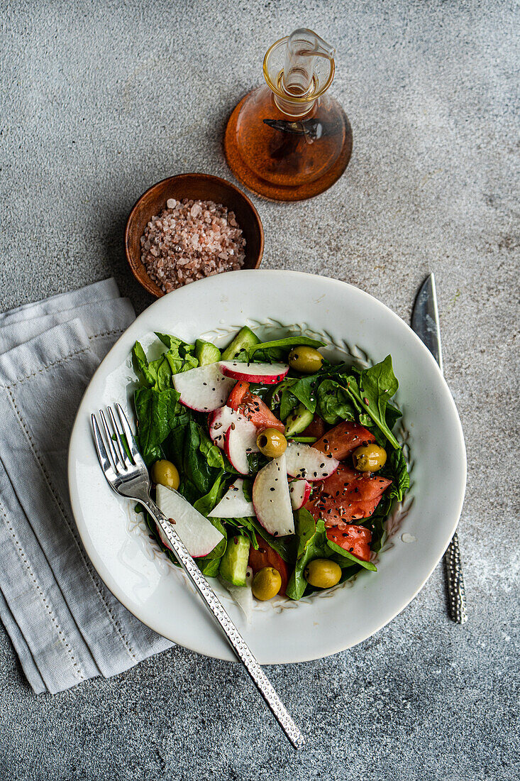 From above of vegetable salad featuring organic spinach leaves, ripe tomatoes, crisp radishes, and green olives beautifully presented in a white bowl with a fork set against a muted backdrop