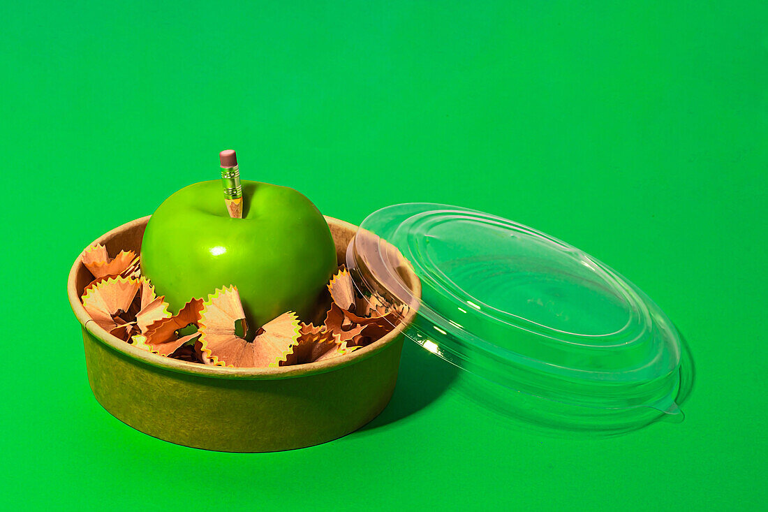 Gesunder Apfel von oben, umgeben von Bleistiftspänen in einer Lunchbox auf grünem Hintergrund, die das Konzept des Null-Abfalls repräsentiert