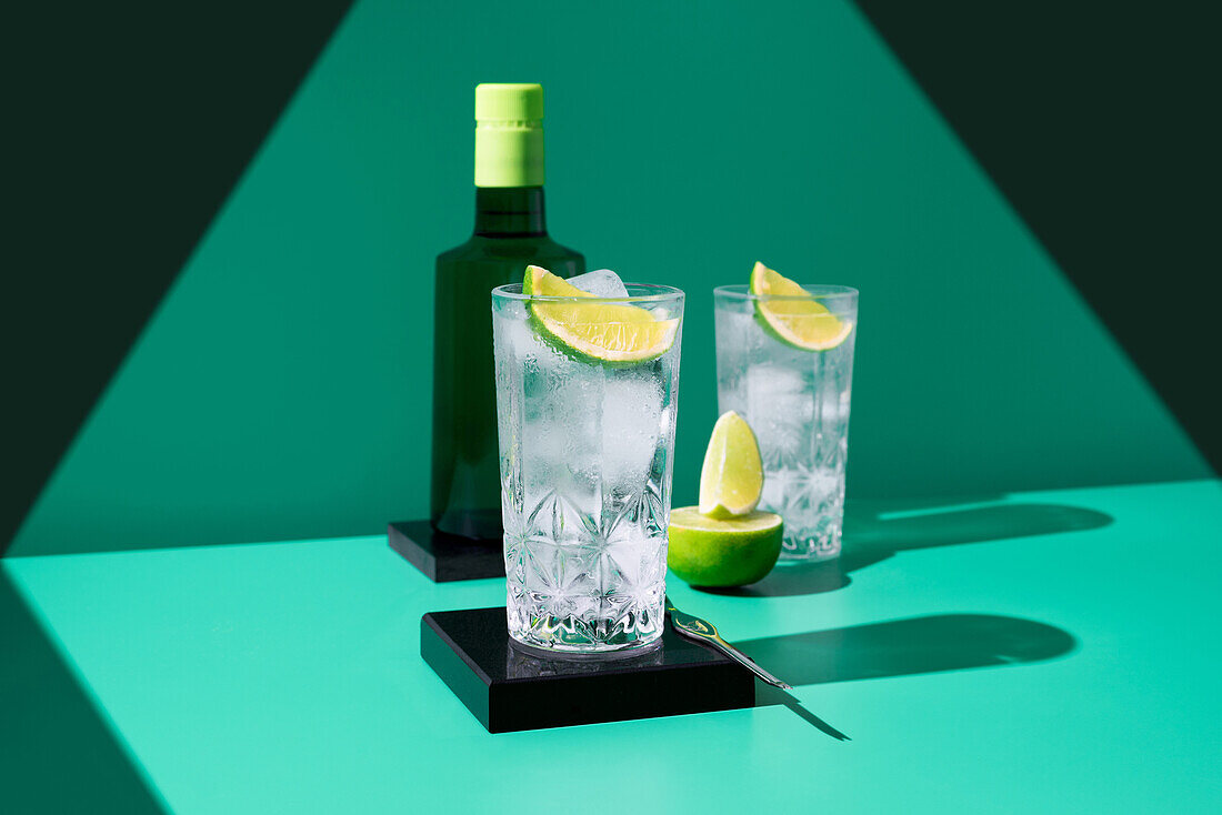 A sophisticated gin tonic presentation featuring two glasses with ice and lime, accompanied by a bottle, on a contrasting teal background