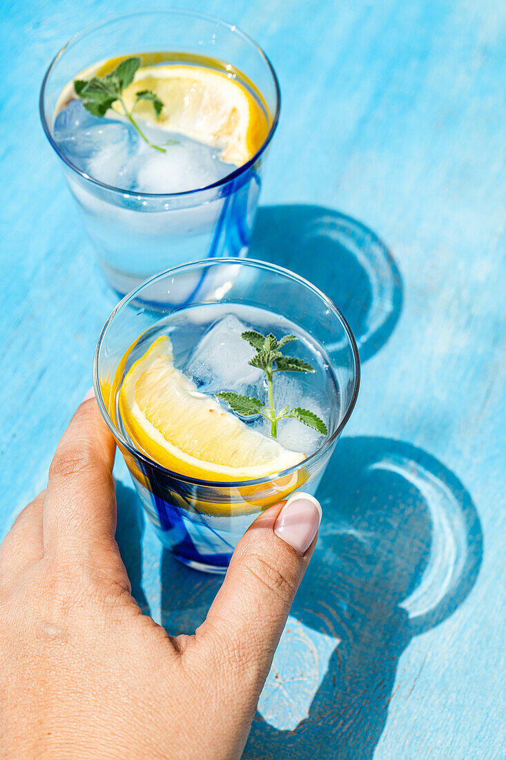 Draufsicht auf eine anonyme Person, die einen Sommercocktail mit Zitronenwodka, Zitronenscheiben und wilden Minzblättern auf einem blauen Tisch mit Eis serviert
