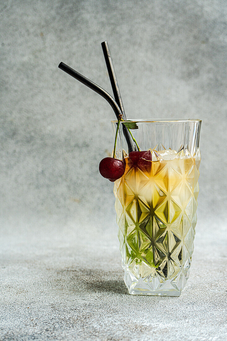 Glass of cold refreshing summer drink with cherry and rosemary sprig placed on gray surface against light background during summer celebration