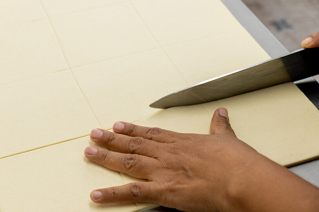 From above of crop anonymous African American baker using knife while cutting smooth sheet of flour dough over table in bakery