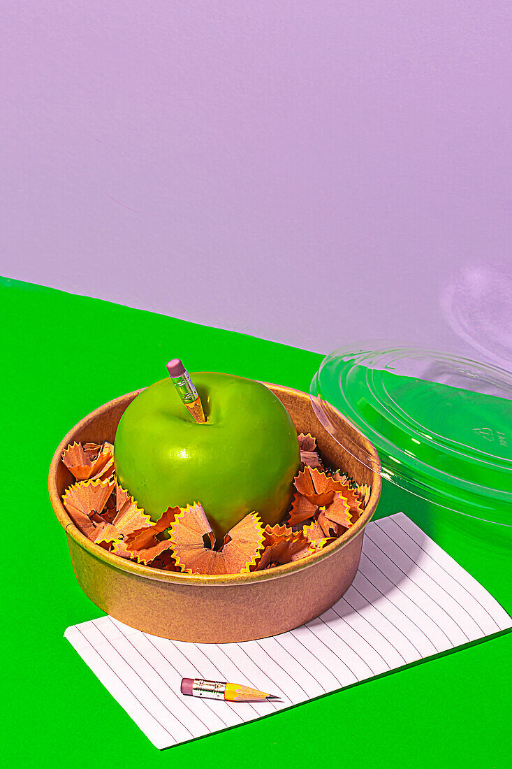 From above of healthy apple surrounded with pencil shavings in lunch box near paper on green table