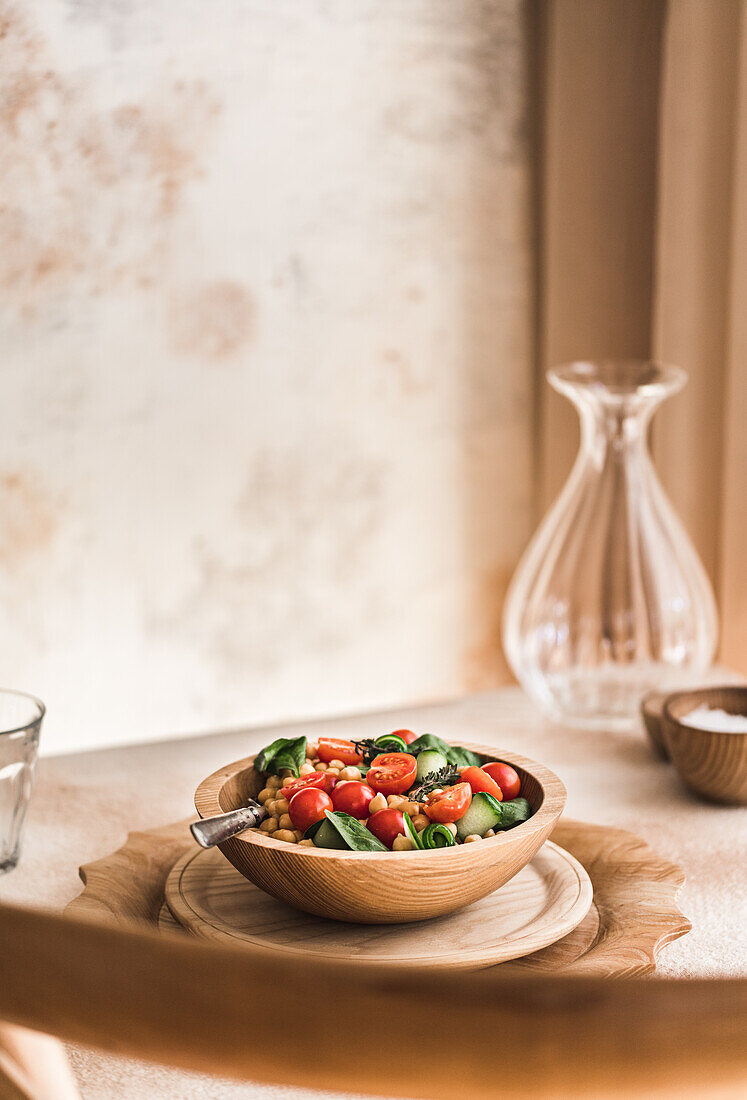 Wooden bowl of delicious salad with fresh vegetables placed on table against glass vase in light kitchen