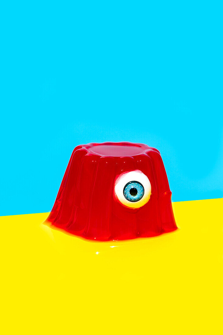 Eyeball with blue iris in red colored jelly pudding placed against vivid blue and yellow backdrop
