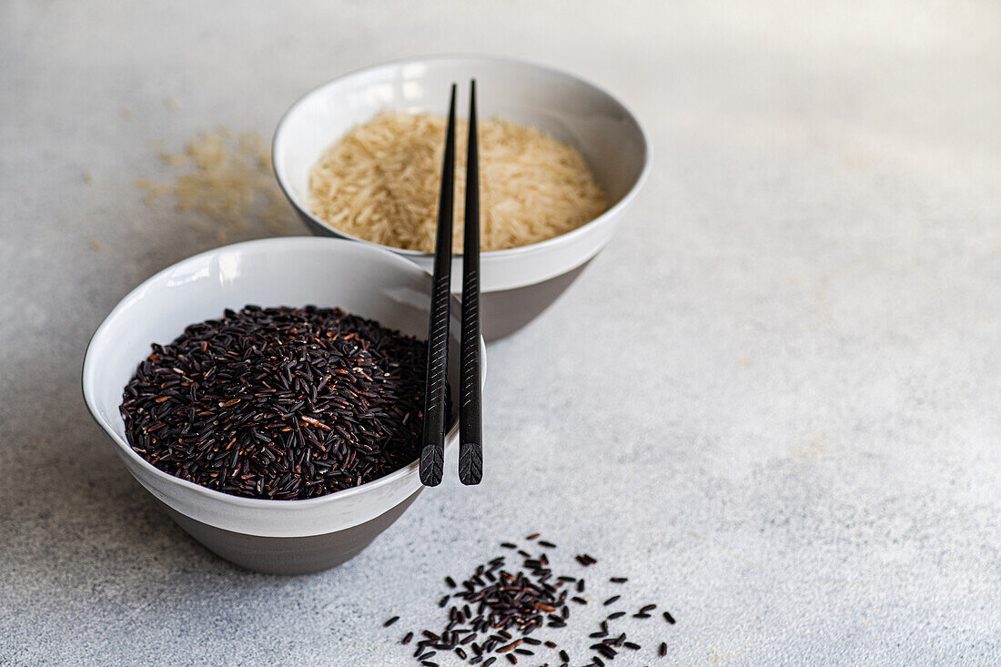 High angle of Raw wild black rice and peeled white rice in the bowls with chopsticks against white surface