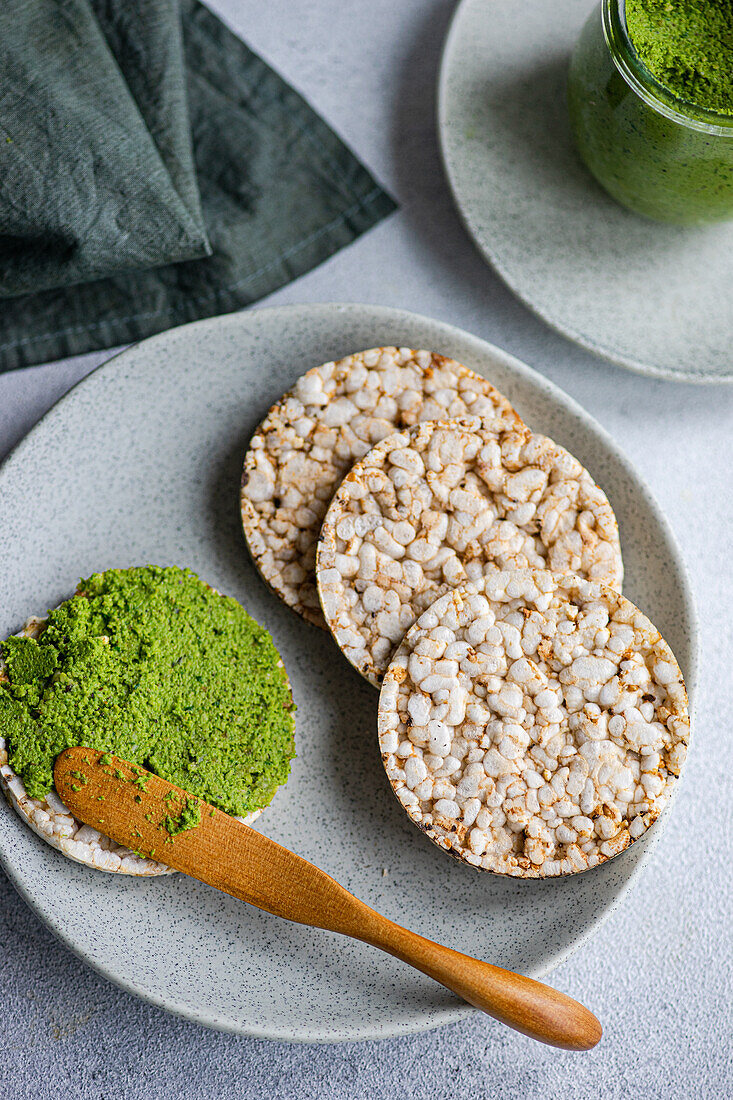 Top view of rice bread on a plate accompanied by a vibrant green spinach pesto pasta-sauce in a glass jar, set against backdrop near napkin