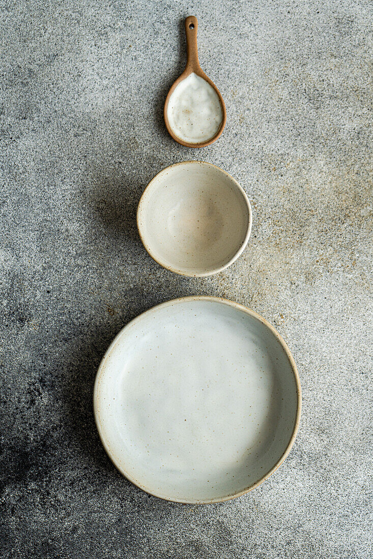 Draufsicht auf Keramikgeschirr, bestehend aus Schüssel, Teller und Holzlöffel, auf einer grauen Fläche