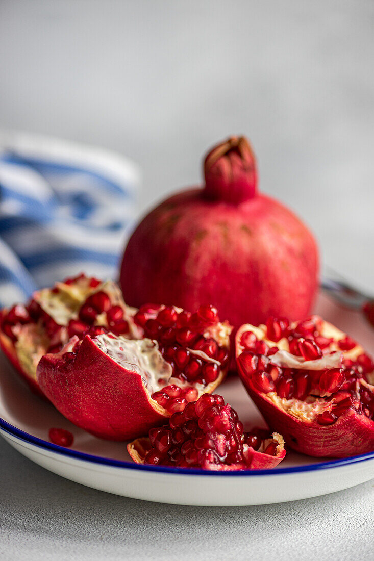 Nahaufnahme von reifen roten Granatapfelkernen auf einem weißen, rustikalen Teller mit blauem Rand auf grauem, strukturiertem Hintergrund