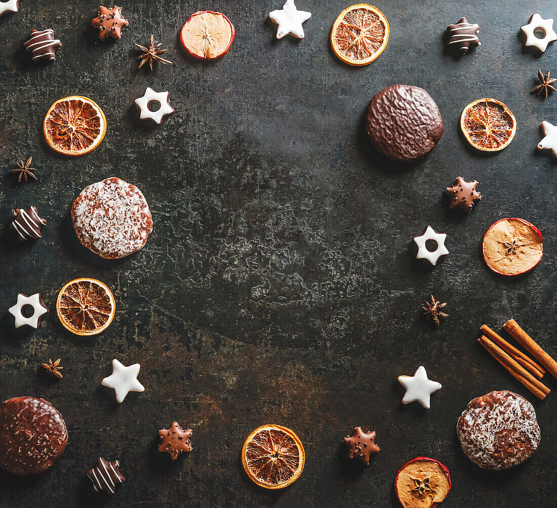 Rahmen mit saisonalen Weihnachtssüßigkeiten und aromatischen Gewürzen: Plätzchen und Lebkuchen, getrocknete Orangenscheiben, Zimt und Sternanis auf dunklem Hintergrund. Draufsicht mit Kopierbereich. Flachlage