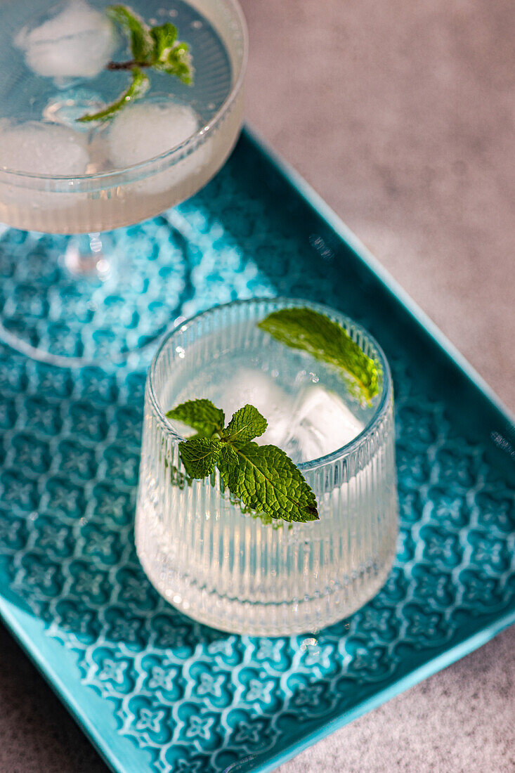 Two refreshing cocktails with mint garnish, ice cubes on a blue patterned tray, elegant glassware