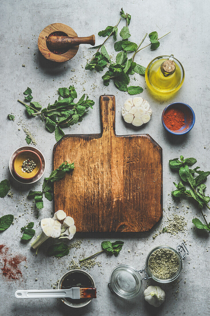 Top view of empty wooden cutting board on grey concrete kitchen table with herbs ,spices, garlic,oil and mortar and pestle