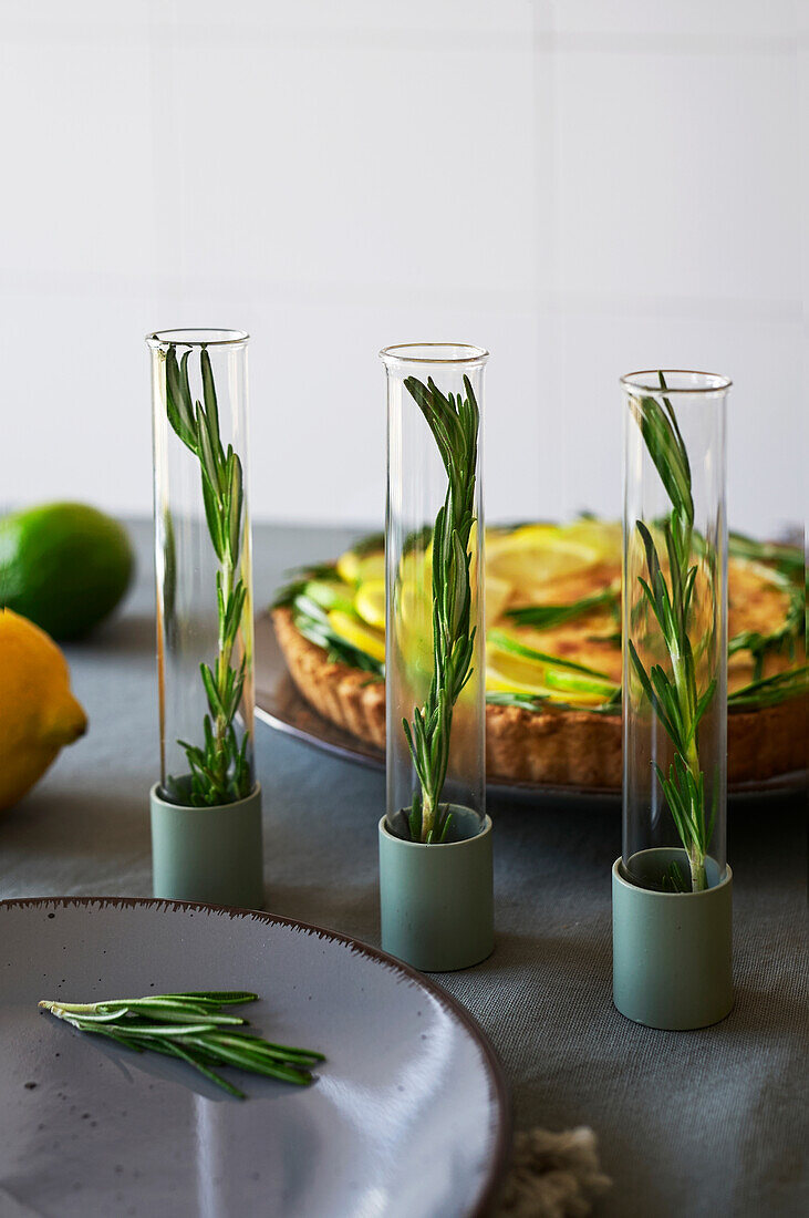 Rosemary leaf placed in a row on a table with a lemon tart in a kitchen against a blurred background