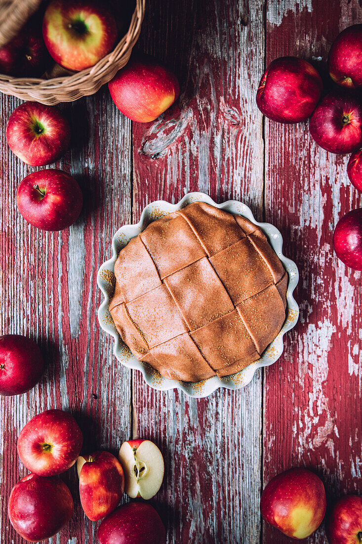 Blick von oben auf einen appetitlichen Bratapfelkuchen, der auf einem schäbigen Holztisch zwischen roten, reifen Äpfeln steht