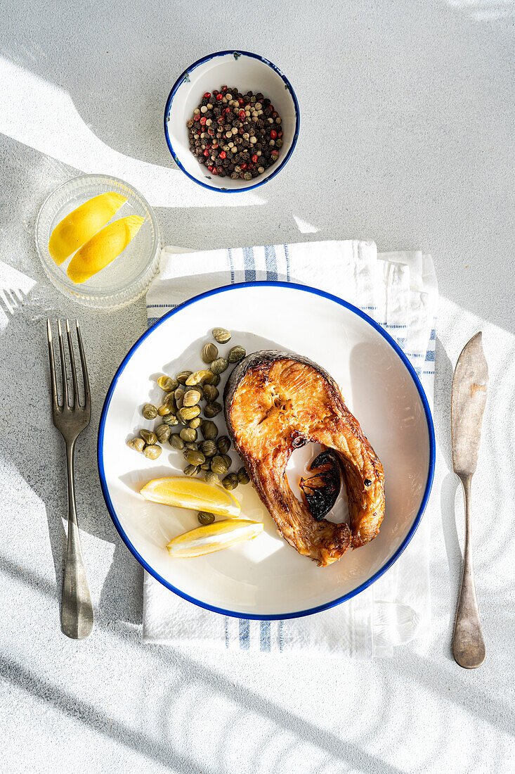 Top view of well-prepared grilled trout steak with capers and lemon served on a white plate with a blue rim placed on striped napkin, cutlery, glass of water and bowl with capers