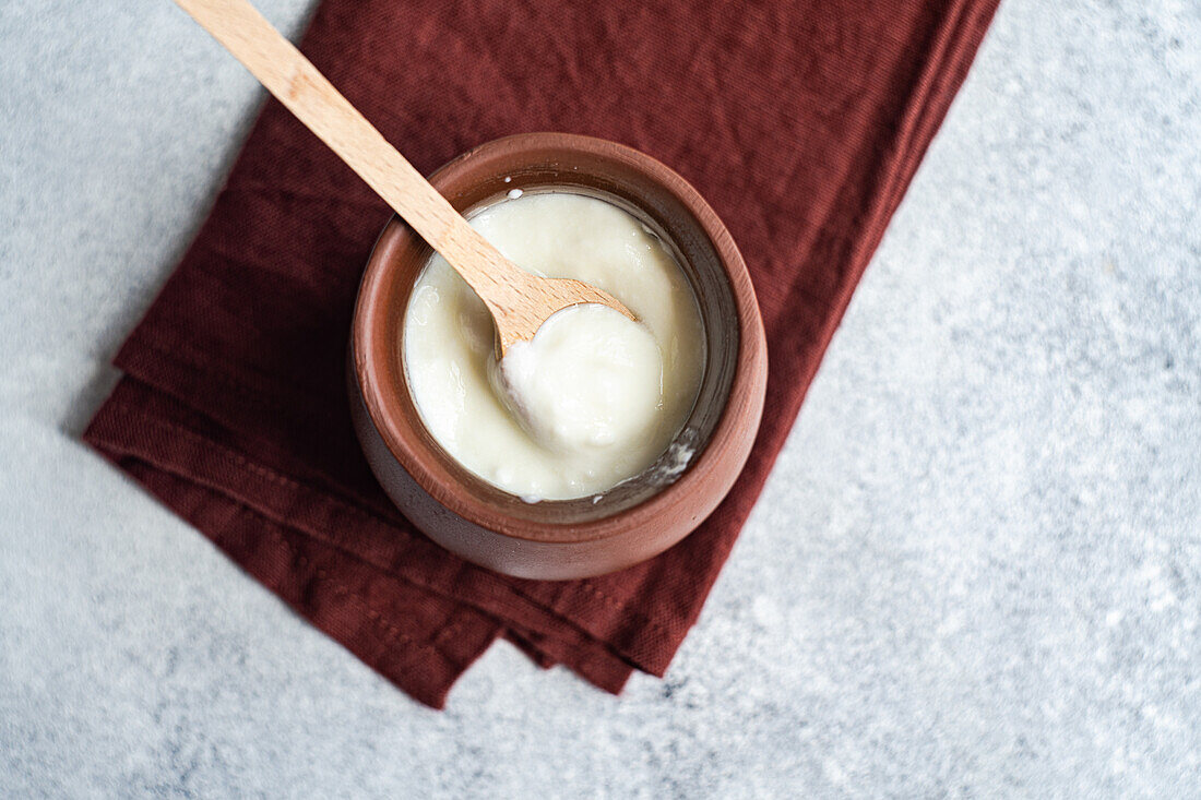 Top view of traditional serving of Georgian sour yogurt known as Matsoni in clay pot with wooden spoon on brown napkin against blurred background