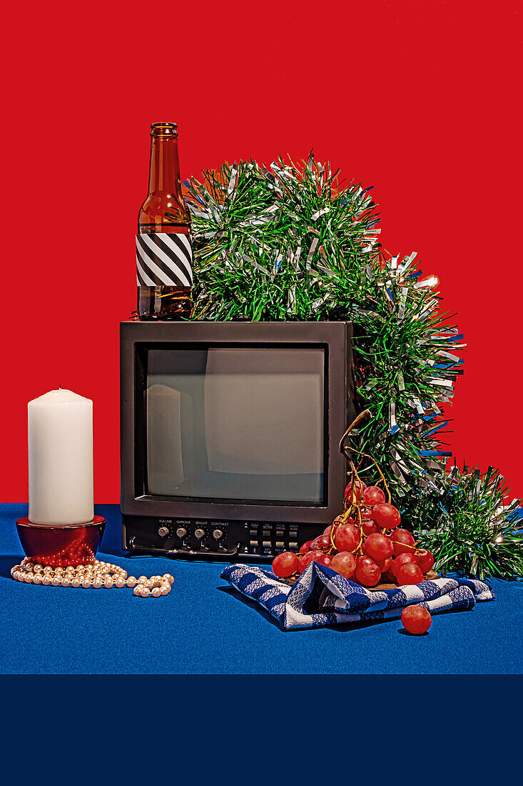 Vintage-Fernsehgerät, umgeben von einer Reihe von Objekten, darunter eine Flasche mit gestreiftem Etikett, frische Trauben auf einem karierten Tuch, eine weiße Kerze und grünes Lametta, alles vor einem roten Hintergrund auf einem blauen Tisch mit einem Gertenfuß mit festlich verzierter Socke