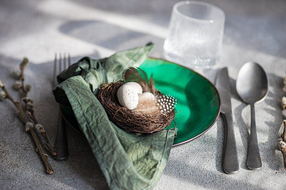 High angle of Easter table setting, showcasing a vibrant green ceramic plate with a small nest with speckled eggs and feathers, placed on gray surface with napkin, cutlery and glass of water