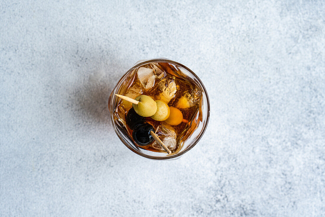 Top view of transparent glass cup with whiskey alcoholic drink and stick of olives cocktail with ice cubes on grey background in studio