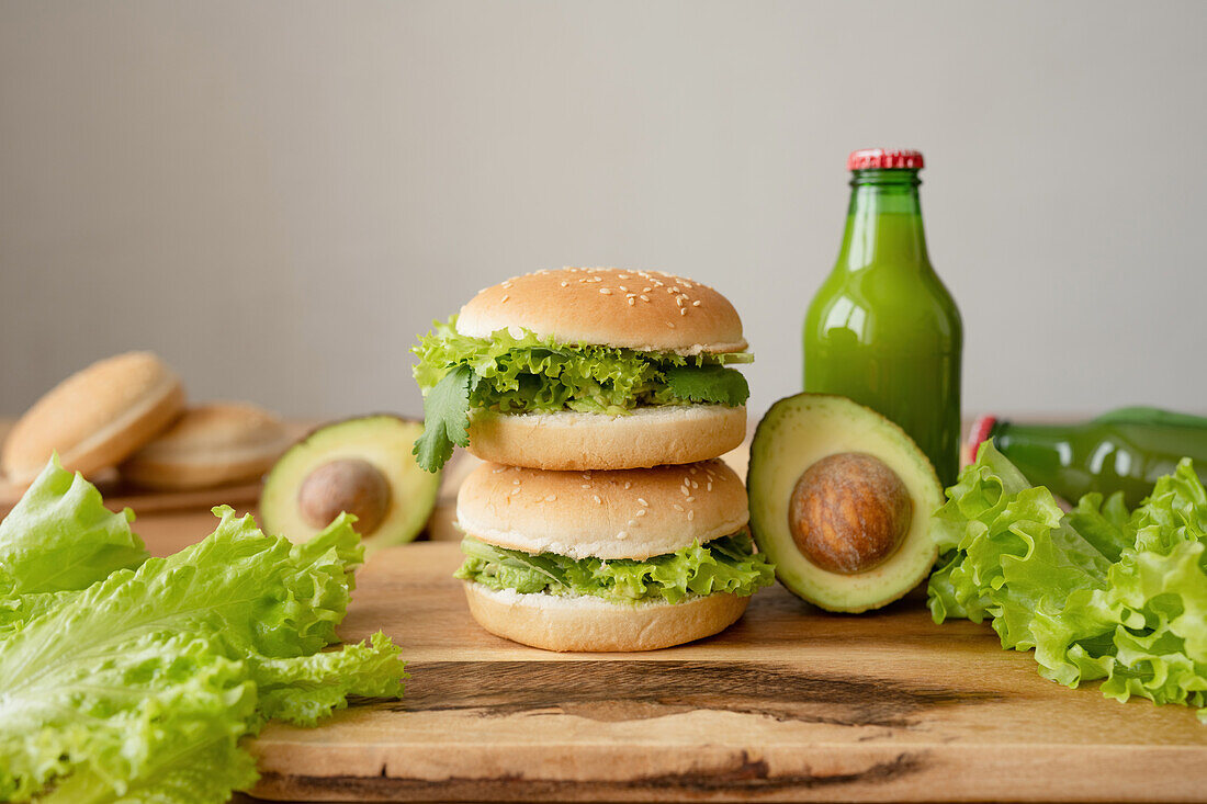 Tasty vegetarian burgers with fresh lettuce near avocado and bottle of juice on wooden cutting board against gray background