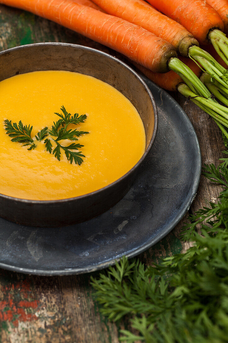A homemade bowl of carrot soup garnished with fresh parsley, surrounded by vibrant carrots on a vintage wooden backdrop.
