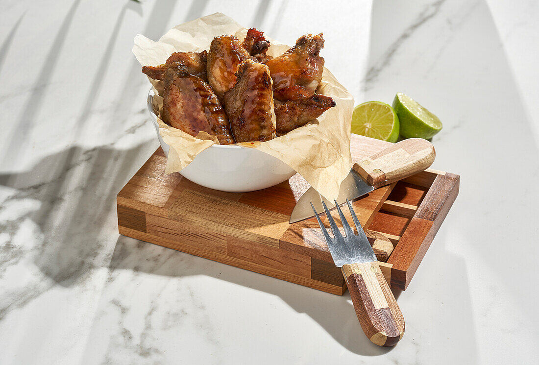 Tasty grilled marinated chicken wings placed in bowl and served with sliced lime on wooden cutting board with knife on table against blurred background in daylight