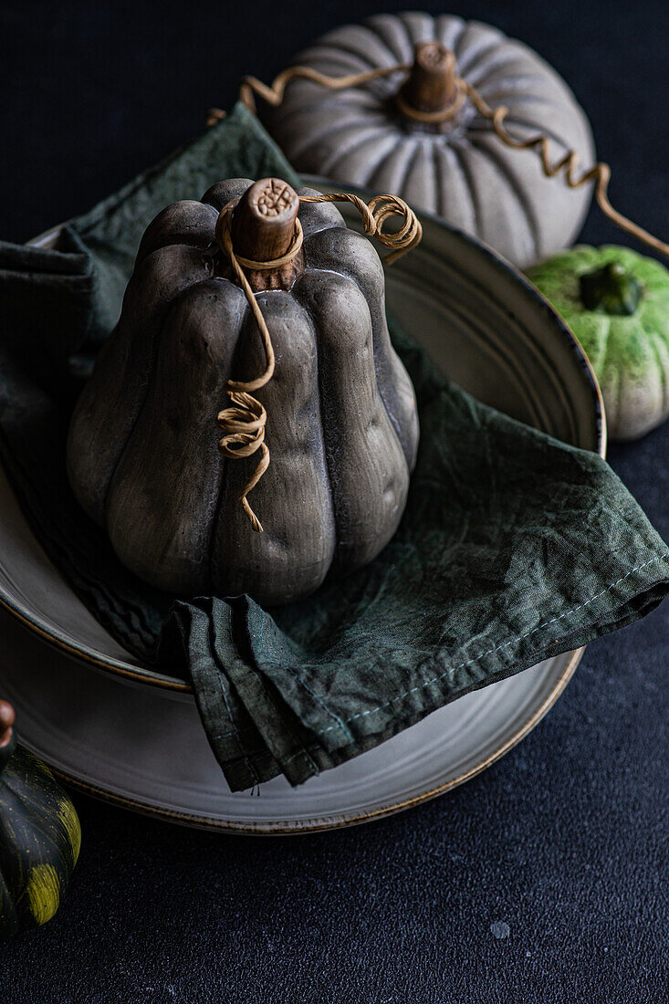 High angle of autumnal table setting with napkin and black pumpkin placed on ceramic bowl near green pumpkins against dark surface