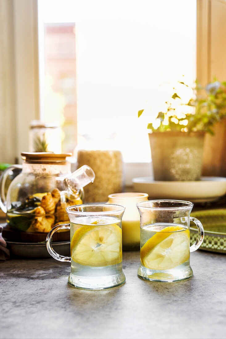 Gesundes Getränk mit Zitrone und Ingwer in gläsernen Teetassen am Küchentisch mit Tassen, Kerze und Kräutern im Fensterhintergrund mit natürlichem Licht. Wärmende Heißgetränke mit Zitrusfrüchten. Vorderansicht