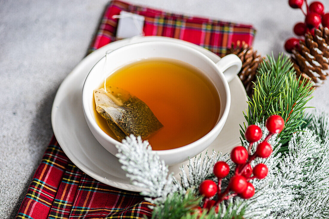 High angle of Christmas cup of tea placed on red napkin near holly and pine cones on gray table